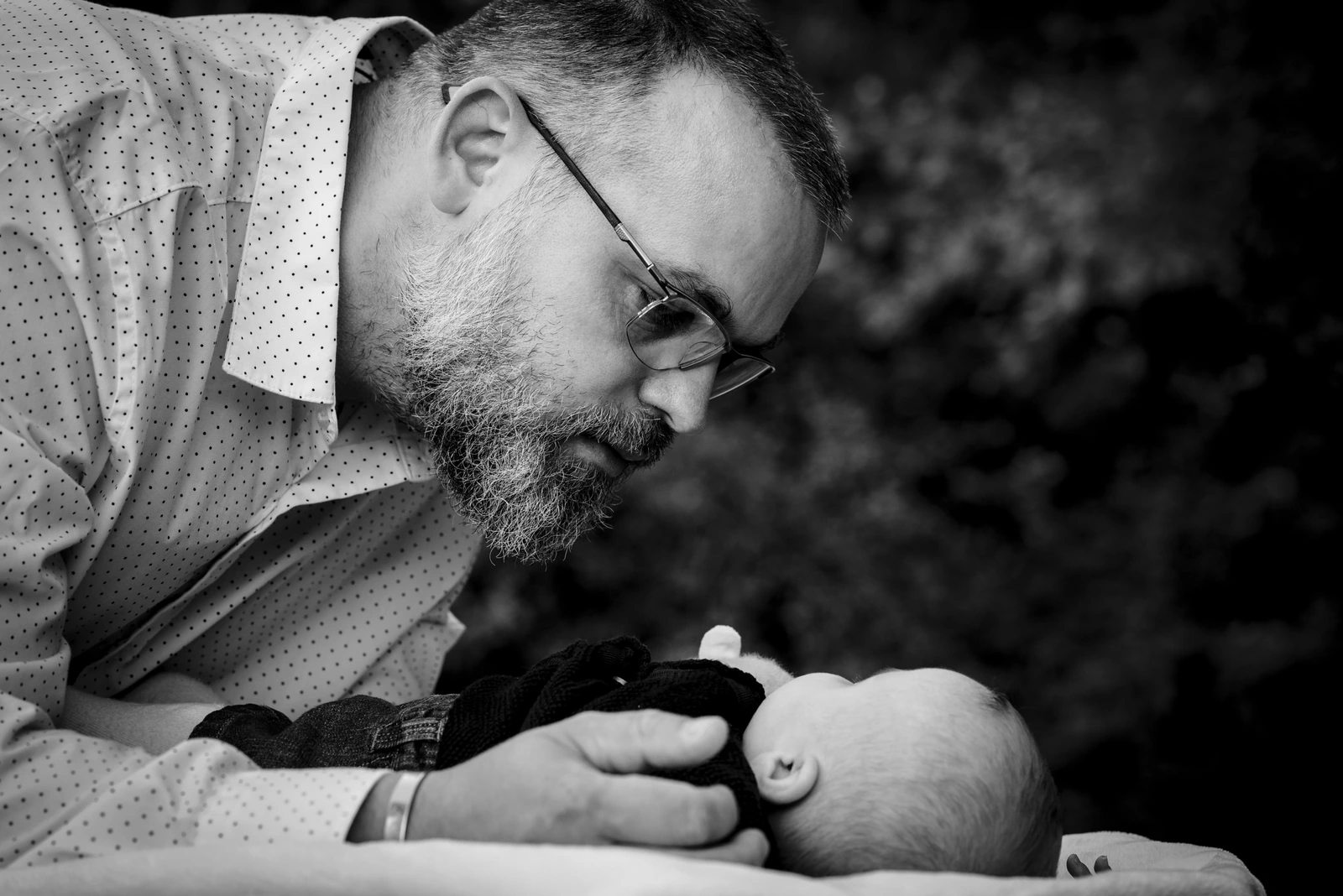 Un homme portant des lunettes se penche vers un bébé allongé, vu de dos, en noir et blanc.